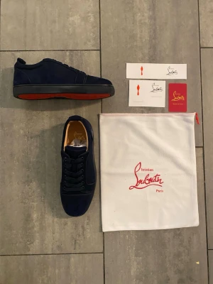 Mörkblå sneakers från Christian Louboutin - Säljer ett par mörkblå sneakers från Christian Louboutin i mocka med den ikoniska röda sulan. Skorna har rund tå, snörning och svarta detaljer. Kommer med originalpåse och äkthetskort. Perfekta för dig som vill sticka ut med lyxig streetstyle.