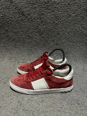 Valentino Flycrews - Säljer ett par Valentino Garavani sneakers i röd mocka med vita skinnpaneler och vita sulor. Skorna har röda snören och en klassisk låg siluett. Märkeslogga på plösen och cool kontrast mellan mocka och skinn. Perfekta för dig som vill sticka ut med stil.