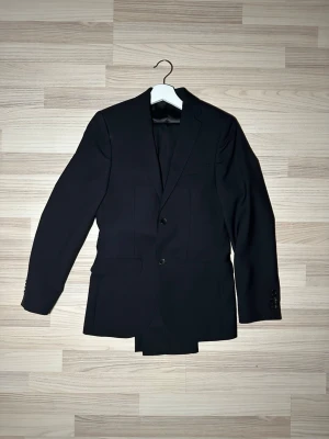 Svart kostym från Riley - Snygg svart kostym från Riley med klassisk passform och två knappar framtill. Kavajen har slät yta, helsvart foder och flera innerfickor. Perfekt för dig som vill ha en stilren och tidlös look. Materialet är ullblandning som känns lyxigt och bekvämt.