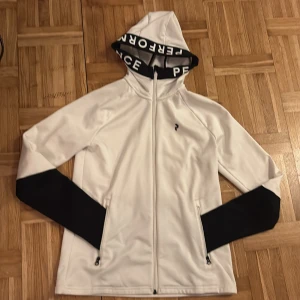 Vit och svart hoodiejacka Peak Performance - Snygg vit hoodiejacka från Peak Performance med svarta detaljer på ärmarna och text på luvan. Jackan har hel dragkedja, två fickor med dragkedja och är tillverkad i mjukt syntetmaterial. Perfekt för dig som gillar sportig och clean stil.