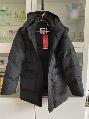 Svart dunjacka från Levi's XS herrstorlek - Säljer en svart dunjacka från Levi's i storlek XS. Jackan är vadderad, har huva och stora fickor framtill. Tillverkad av 100% återvunnen polyester och är vattenavvisande. Perfekt för kalla dagar och har dragkedja framtill. Oanvänd med lappar kvar.