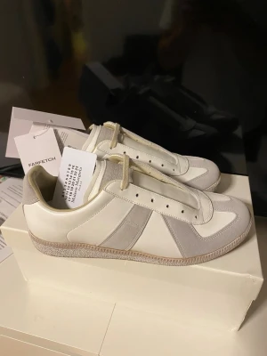Maison Margiela Gats sneakers vit/grå - Maison Margiela Gats sneakers i vitt och ljusgrått med klassisk retrodesign. Skorna har ovandel i skinn och mocka, beige snörning och gummisula med präglad logga undertill. Perfekt för dig som gillar minimalistisk och stilren vibe.