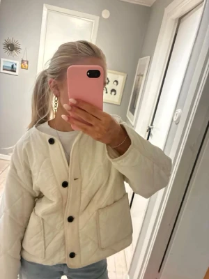 Quiltad vit jacka med knappar - Säljer en stilren quiltad vit jacka med svarta knappar och stora fickor framtill. Jackan har en boxig passform och är perfekt för dig som gillar en clean och modern look. Passar snyggt till jeans eller kjol och är lätt att matcha med olika outfits.