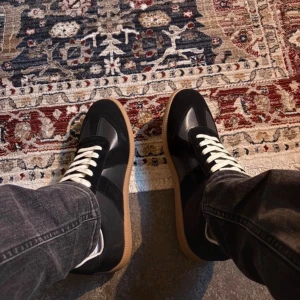 Maison Margiela svarta sneakers i skinn - Maison Margiela sneakers i svart skinn och mocka med beige snörning och klassisk gummisula. Snygg kontrast mellan material och färger, perfekt till jeans eller streetwear. Ikonisk design med rund tå och platt sula. Märkeslogga under sulan.