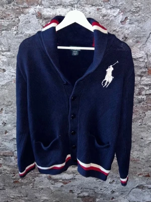 Marinblå kofta från Polo Ralph Lauren - Säljer en marinblå kofta från Polo Ralph Lauren med stora vita loggan broderad på bröstet. Sitter som en M! Koftan har knappar framtill, två fickor och ribbade muddar med röda och vita ränder. Snygg krage med samma randiga detaljer. Perfekt till höst och vår. Några frågor kom DM!😆