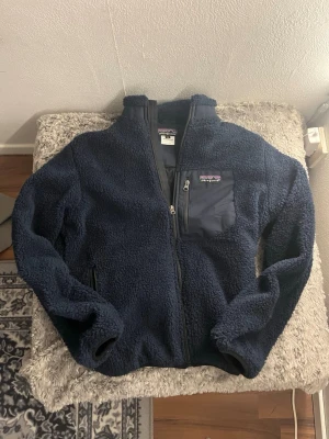Mörkblå fleecejacka från Patagonia - Säljer en mörkblå fleecejacka från Patagonia med hel dragkedja och bröstficka med dragkedja. Jackan är fluffig och har hög krage samt två sidofickor. Perfekt för kyliga dagar och outdoor vibes.
