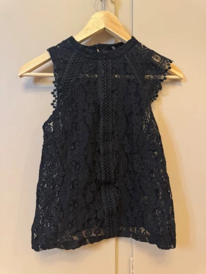 Svart spetsblus från Primark - Säljer en svart ärmlös spetsblus från Primark i storlek 38. Blusen har hög hals, vackra spetsdetaljer och dekorativa kanter med små bollar vid axlarna. Perfekt för dig som gillar en feminin och elegant look.