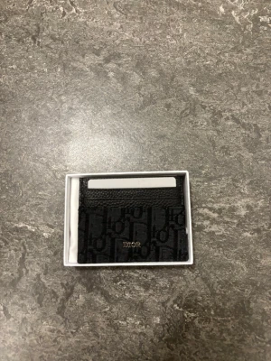 Svart  plånbok i skinn och textil - Säljer en svart  plånbok i smidig modell med klassiskt mönster. Plånboken har flera kortfack och är tillverkad i en mix av skinn och textil. Kommer med originalbox, dustbag och äkthetskort. Perfekt för dig som vill ha något stilrent och lyxigt.