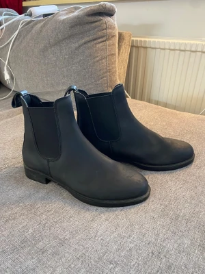 Svarta chelsea boots i skinn, storlek 39 - Säljer ett par klassiska svarta chelsea boots i skinn med elastiska paneler på sidorna och dragflik bak. Bootsens sula är platt och har ett diskret mönster för bra grepp. Perfekta till höst- och vinterlooken.