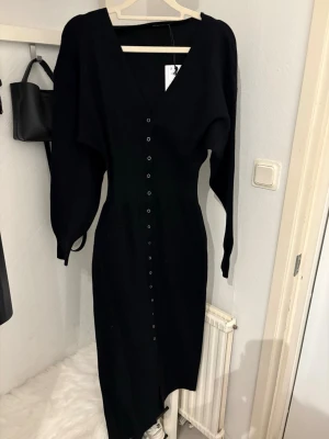Svart lång stickad klänning - Säljer en svart lång stickad klänning med v-ringning och knappar längs hela framsidan. Klänningen har långa ärmar och en smickrande passform som markerar midjan. Perfekt för dig som gillar stilrena och bekväma plagg.      Klänningen är från Gina Tricot och är endast testad, prislappen är på. Inget fel på klänningen.