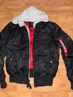 Svart bomberjacka från Alpha Industries - Säljer en svart bomberjacka från Alpha Industries med grå huva och röd innerfoder. Jackan har dragkedja, ribbade muddar, fickor och en ikonisk röd tag på ärmen. Perfekt för dig som vill ha en streetstyle-look.