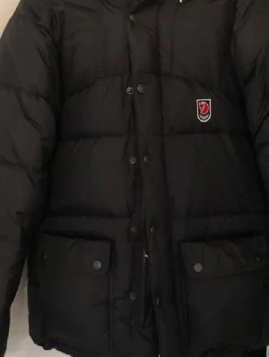 Svart dunjacka från Fjällräven - Säljer en svart dunjacka från Fjällräven med huva och stora fickor framtill. Jackan har dragkedja och knappar, samt den klassiska Fjällräven-loggan på bröstet. Perfekt för kalla dagar och riktigt skön att bära. Har mycket hål samt tappat dun