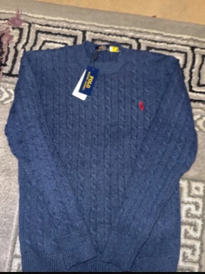 Blå kabelstickad tröja Polo Ralph Lauren - Kabelstickad blå tröja från Polo Ralph Lauren med rund halsringning och den klassiska röda logotypen broderad på bröstet. Tröjan har långa ärmar och ribbade muddar vid ärmslut och nederkant. Perfekt för dig som gillar stilren och tidlös design.
