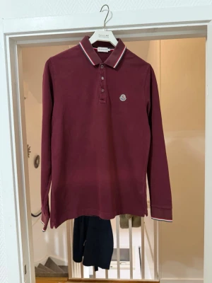 Vinröd långärmad pike Moncler - Säljer en vinröd långärmad henley från Moncler med klassisk krage och knappar framtill. Piken har vita och marinblå detaljer på krage och ärmslut samt Moncler-logga på bröstet. Tillverkad i mjuk bomull som känns skön mot huden. Og tags