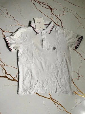 Vit Moncler pikétröja med ränder - Vit pikétröja från Moncler med klassisk krage och knäppning framtill. Kragen och ärmsluten har tunna ränder i rött och marinblått. Tröjan har korta ärmar och Moncler-logga på bröstet. Perfekt för en clean och sportig look.