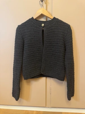 Mörkgrå stickad kofta från Zara - Mysig mörkgrå stickad kofta från Zara med rund halsringning och ett dekorativt knappslå framtill. Koftan har lång ärm och ett strukturerat, ribbat mönster som ger en cool och modern vibe. Perfekt att slänga över en t-shirt eller topp.