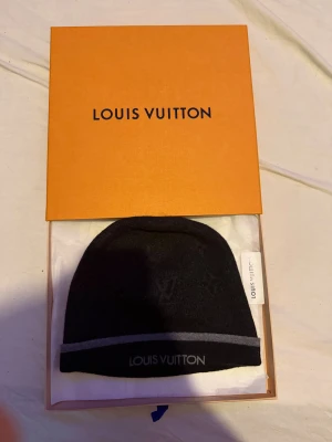 Louis Vuitton svart mössa med logga - Svart mössa från Louis Vuitton med diskret LV-monogram och grå rand längs kanten. Snyggt broderad logotyp framtill. Kommer i originalförpackning med kvitto. Perfekt accessoar för dig som gillar exklusiva märken och stilren design.