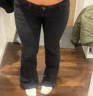 Svarta bootcut jeans med fickdetaljer - Snygga svarta jeans med bootcut passform och klassiska fem fickor, varav bakfickorna har lock och silverfärgade knappar. Jeansen har låg midja och är tillverkade i ett stretchigt denimtyg som sitter bekvämt och följer kroppen. Lite slitage i byxkanten annars fint skick