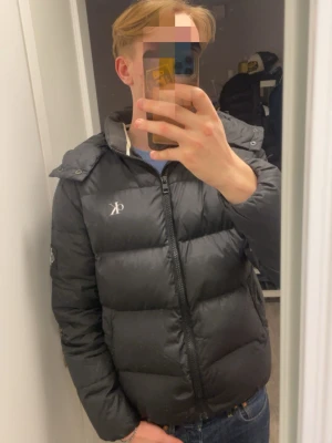 Svart pufferjacka från Calvin Klein - Säljer en svart pufferjacka från Calvin Klein Jeans med huva och dragkedja framtill. Jackan har CK-logga på bröstet och ärmen, quiltad design och lång ärm. Perfekt för kalla dagar och har en clean, modern look som funkar till jeans och sneakers.