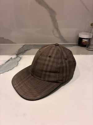 Brun rutig Burberry keps i bomull - Snygg brun keps från Burberry med klassiskt rutigt mönster och broderad logga baktill. Kepsen är tillverkad i 100% bomull med detaljer i lammnappa och har justerbar rem bak. Perfekt accessoar för en cool och stilren look.