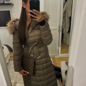 Lång beige dunjacka med pälshuva - Säljer en lång, quiltad beige dunjacka med stor fluffig pälskantad huva och knytskärp i midjan. Jackan har knappar framtill och långa ärmar med svarta muddar. Perfekt för kalla dagar och riktigt snygg siluett.