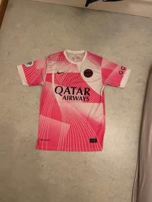 PSG Mbappé matchtröja rosa Nike - Säljer en Paris Saint-Germain matchtröja med Mbappé 7 på ryggen. Tröjan är designad i rosa och vitt med grafiskt mönster och svarta detaljer. PSG-logga på bröstet och Qatar Airways som sponsor. Tillverkad i lätt polyester för bästa komfort.