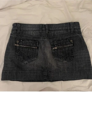 Svart jeanskjol med dragkedjor från YFG - Cool svart jeanskjol från YFG Jeans med snygga detaljer som dragkedjor på fickorna och slitningar för en edgy look. Kjolen har klassisk femficksdesign, bälteshällor och knappstängning framtill. Perfekt för dig som gillar streetstyle och denim.