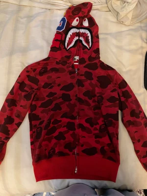 BAPE Shark Hoodie – Röd Camo (Storlek S) - Säljer en röd A Bathing Ape (BAPE) Shark Hoodie i storlek S. Hoodien har det klassiska camo-mönstret och hel zip-design med haj-huvan. Passar både tjejer och killar (unisex).
