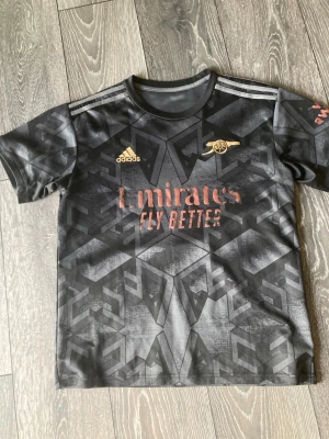 Arsenal tränings T-shirt  - svart och grå Arsenal fotbollströja från Adidas med geometriskt mönster och bronsfärgade tryck