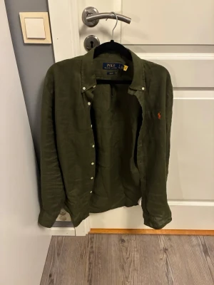 Olivgrön linneskjorta Polo Ralph Lauren - Olivgrön skjorta från Polo Ralph Lauren i slim fit med klassisk krage och vita knappar. Tillverkad i linne, med den ikoniska orange broderade loggan på bröstet. Långärmad och stilren, perfekt för dig som gillar clean och chill look.