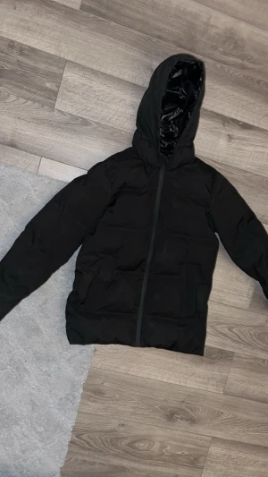 Svart pufferjacka från Jack & Jones - Svart pufferjacka från Jack & Jones med huva och hel dragkedja framtill. Jackan har två stora fickor och är tillverkad i syntetmaterial med glansig insida. Perfekt för kalla dagar och har en clean, stilren look.