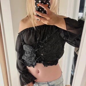 Svart offshoulder croppad blus - Säljer en svart offshoulder blus med croppad passform och långa, transparenta ärmar. Toppen har snygg snörning framtill och broderade detaljer i tyget. Perfekt för dig som gillar edgy och romantisk stil. Passar till både jeans och kjol. Vet ej storlek men ungefär xs/s