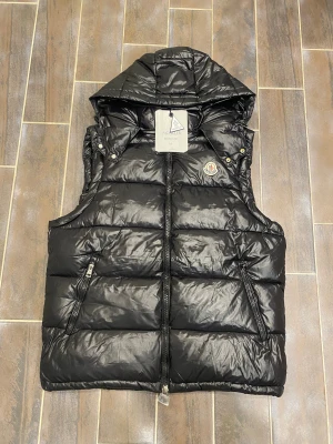 Moncler väst - Svart dunväst från Moncler med avtagbar huva och klassisk logotyp på bröstet. Västen har quiltad design, två fickor med dragkedja och snygga guldfärgade tryckknappar vid axlarna. Insidan har ett coolt serietidningsmotiv. Perfekt för lager-på-lager under höst och vinter. NFC funkar.