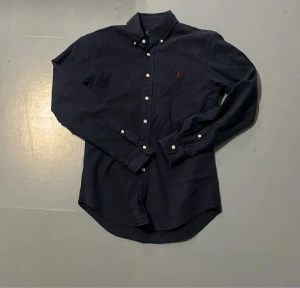 Mörkblå skjorta från Ralph Lauren - Snygg mörkblå skjorta från Ralph Lauren med klassisk button-down krage och vita knappar. Skjortan har lång ärm och den ikoniska röda logon broderad på bröstet. Perfekt för dig som gillar stilrena och tidlösa plagg.