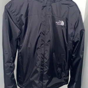 The North Face storlek S inte L!!! Vattentätt och vindtätt  - Svart vindjacka från The North Face med huva och dragkedja framtill. Jackan har logga på bröstet och ryggen, samt innerfoder med märkets band. Materialet är syntet och polyester, perfekt för blåsiga dagar. Snygg och funktionell design med långärmade ärmar.                                                              Storlek S inte L!!!                                                                 Vattentätt och vindtätt