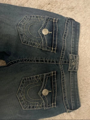 Blå bootcut jeans från True Religion - Snygga blå jeans från True Religion med bootcut passform och klassiska kontrastsömmar. Baksidan har två fickor med lock och silverfärgade knappar samt märkets logga på linningen. Jeansen har låg midja och är tillverkade i jeansmaterial.