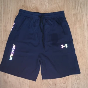 Marinblå träningsshorts från Under Armour - Snygga marinblå träningsshorts från Under Armour med reflekterande logga och text på benet. Shortsen har elastisk midja med snörning och är tillverkade i ett lätt och snabbtorkande syntetmaterial, perfekt för träning eller sportiga dagar. Ena dragkedjan är lite trög och själva grejen man drar med har lossnat