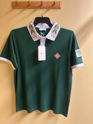 Grön polotopp från Casablanca - Casablanca Tennis Club polotopp i mörkgrönt med vit krage och ärmslut. Kragen har broderade detaljer i grönt och guld. Patch med Casablanca-logga på ärmen och röd emblem på bröstet. Topp i bomull med korta ärmar, perfekt för en sportig vibe.