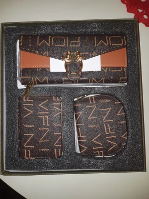 Brun plånboksset från Vivixen - Snyggt set med brun plånbok, kortfodral och myntbörs från Vivixen. Alla delar har ett coolt grafiskt mönster i beige och brunt, samt guldfärgade detaljer. Plånboken har en unik dekoration med två fåglar på framsidan och stilren design.