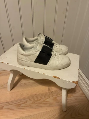 Valentino Open sneakers vit/svart - Snygga Valentino Open sneakers i vitt skinn med bred svart rem över snörningen. Klassisk rund tå och platt sula, med diskret logga på plösen. Perfekt för dig som gillar stilrena och lyxiga sneakers med en unik touch.