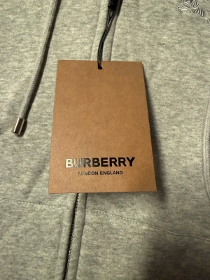 Grå hoodie från Burberry med huva - Snygg grå hoodie från Burberry med klassisk broderad ryttare på bröstet. Tröjan har huva med snörning, dragkedja framtill och ribbade muddar. Insidan av huvan har Burberrys ikoniska rutmönster. Tillverkad i mjuk bomull för skön komfort.