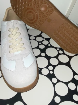 Maison margiela - Snygga sneakers i vitt med beige mockadetaljer och krämfärgade snören. Skorna har en klassisk rund tå och platt gummisula i brunt. Materialmixen av skinn och mocka ger en fräsch och stilren look som passar perfekt till jeans eller kjol.