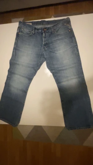 Blå bootcut jeans från Jack & Jones - Säljer ett par klassiska blå jeans från Jack & Jones Vintage Denim. De har en rak passform, fem fickor och snygga slitningar framtill. Jeansen är tillverkade i jeansmaterial och har gul kontrastsöm vid midjan. Perfekt för dig som gillar en enkel och tidlös stil.