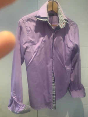 Lila skjorta från Make Love not War - Snygg lila skjorta med lång ärm och coola svartvita ränder på insidan av krage och manschetter. Skjortan har klassisk krage och knappar framtill. Perfekt för dig som vill sticka ut med färg och detaljer. Materialet känns som bomull och passformen är normal.