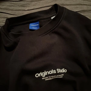 Svart sweatshirt från Jack & Jones XS - Svart sweatshirt från Jack & Jones Originals Studio i storlek XS. Tröjan har ett stilrent tryck på bröstet och är tillverkad i mjuk bomull med långärmad passform. Perfekt för dig som gillar enkel och clean streetstyle.