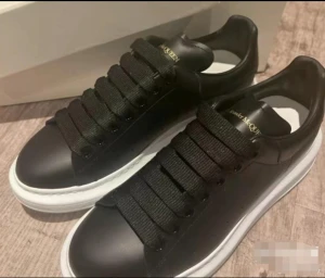 Svarta sneakers Alexander McQueen - Snygga svarta sneakers från Alexander McQueen med breda svarta snören och vit platt sula. Skorna har en stilren design med diskret guldfärgad logga på plösen. Perfekta för dig som gillar chunky och trendiga sneakers.