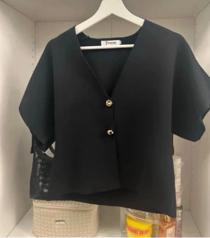 Svart blus - Svart blus med korta ärmar och två snygga guldfärgade knappar framtill. Blusen har en v-ringad design och är lite boxig i modellen, perfekt att styla över en t-shirt eller linne. Enkel och stilren look som funkar till många outfits.