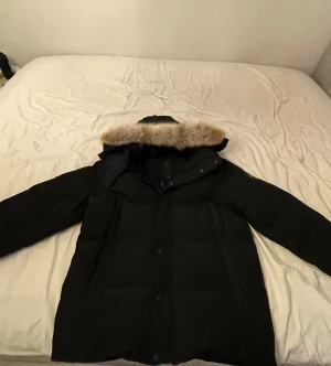 Svart  Canada Goose black badge  - Svart dunjacka från Canada Goose i storlek XL. Jackan har en huva med beige pälsdetalj, klassisk patch på ärmen och flera fickor framtill. Perfekt för kalla vinterdagar med sin långa passform och varma material. Snygg och tidlös design som håller dig varm.