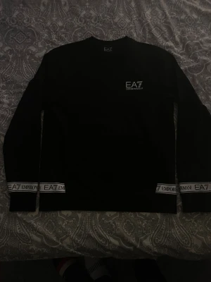 Svart långärmad EA7 Emporio Armani tröja - Svart långärmad tröja från EA7 Emporio Armani med vit logga på bröstet och snygga vita band med logotyp vid ärmslut och nederkant. Tröjan har en klassisk rund hals och är gjord i mjuk bomull som känns skön mot huden. Perfekt för dig som gillar stilrena och exklusiva plagg.