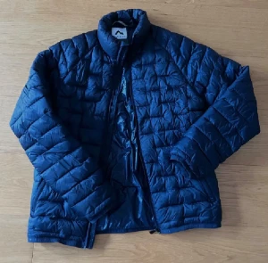 Blå quiltad dunjacka från McKinley - Blå dunjacka från McKinley i quiltad design. Jackan har dragkedja framtill och är lätt vadderad för extra värme. Perfekt för kyliga dagar och enkel att matcha med olika outfits.